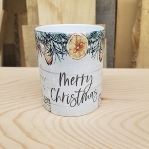 May include: White ceramic mug with a wintery design featuring pine branches, pine cones, and orange slices. The mug has the text "Merry Christmas" printed on the front.