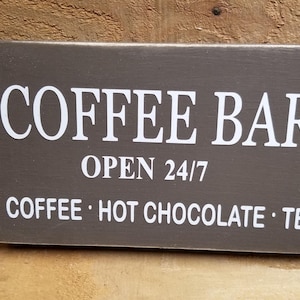 May include: A brown wooden sign with white text that reads "Coffee Bar Open 24/7 Coffee - Hot Chocolate - Tea".