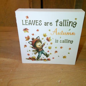 May include: A white rectangular wooden block sign featuring a cartoon illustration of a child wearing a fox-ear hat, scarf, and boots, surrounded by falling autumn leaves. The text reads: LEAVES are falling Autumn is calling.