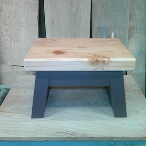 May include: A small wooden stool featuring a rectangular top with a natural light wood grain finish and visible knots. The stool is supported by four sturdy, angled legs painted in a solid dark charcoal gray color, providing a stable base.