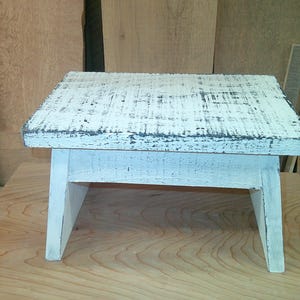 May include: A small wooden step stool featuring a rectangular top and four angled legs. The stool is finished with a distressed white paint application that reveals dark wood grain textures across the entire surface of the piece of furniture.