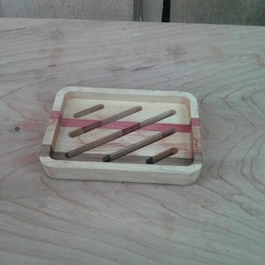 May include: A rectangular wooden soap dish with a light wood frame and a red stripe. The dish has several brown wooden slats for drainage. The soap dish is designed to hold a bar of soap and keep it dry.