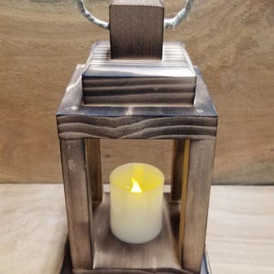 May include: A rustic wooden lantern with a rope handle and a clear glass panel. A white candle is inside the lantern, casting a warm glow.