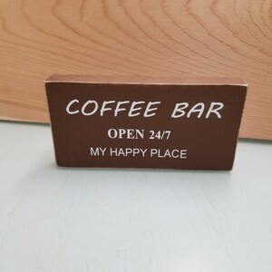 May include: A brown wooden sign with white text that reads "Coffee Bar Open 24/7 My Happy Place".
