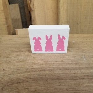 May include: A white wooden block with three pink bunny silhouettes painted on it. Each bunny has a small white heart on its backside.