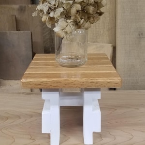 May include: A small white wooden stool with a square top made of light brown wood. A glass vase with dried flowers sits on the top of the stool.