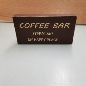 May include: A brown wooden sign with the text "COFFEE BAR" in white lettering. Below the text is "OPEN 24/7" and "MY HAPPY PLACE" in white lettering.