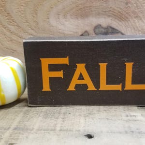 Puede incluir: Un letrero de madera con la palabra "FALL" pintada en letras naranjas sobre un fondo marrón oscuro. Una pequeña calabaza blanca con rayas amarillas está en primer plano.