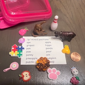 May include: A set of colorful plastic toys for learning phonics. The toys include a puzzle, a pineapple, a paw print, a pear, a penny, a pan, a pin, a pumpkin, an apple, a grape, an ape, a hippo, a puzzle, and a  card with the words '/p/ mixed positions' and a list of words starting with the letter 'p'.