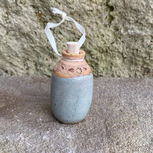 May include: A small, handmade ceramic bottle with a cork stopper and a white ribbon. The bottle has a light blue body and a tan-colored top with decorative cutouts. The bottle is approximately 3 inches tall.