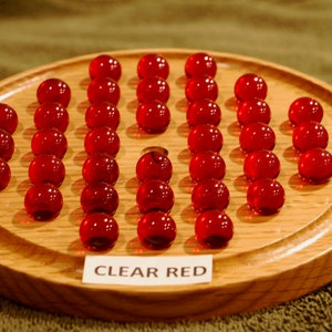 May include: A wooden game board with a circular playing area. The board is filled with 24 clear red glass marbles arranged in a grid pattern. The text "CLEAR RED" is printed on a label at the bottom of the board.