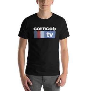 Può includere: T-shirt nera con la scritta "corncob tv" in bianco, sopra un rettangolo orizzontale con strisce blu, rosse e azzurre. La parola "tv" è bianca su sfondo blu.