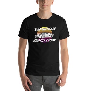 Può includere: T-shirt nero con una grafica di un'auto sportiva bianca e il testo "Dangerous Nights Crew" in un design di tramonto retrò.