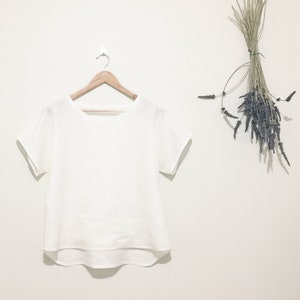 May include: A white, short-sleeved top hangs on a wooden hanger against a white wall. The top has a rounded neckline and a slightly curved hem. Dried lavender and grasses are tied together and hang on the wall to the right.