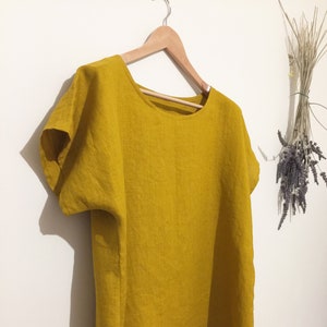 May include: Mustard yellow linen top with short sleeves and a rounded neckline. The top is hanging on a wooden hanger against a white wall. A bundle of dried flowers is visible in the background.