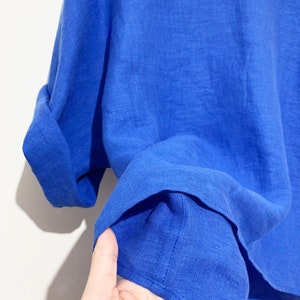 May include: A close-up of a vibrant blue linen top. The short-sleeved shirt has rolled-up cuffs and a round neckline. The fabric appears lightweight and slightly textured, suggesting breathability. The bright color makes it a stylish garment.
