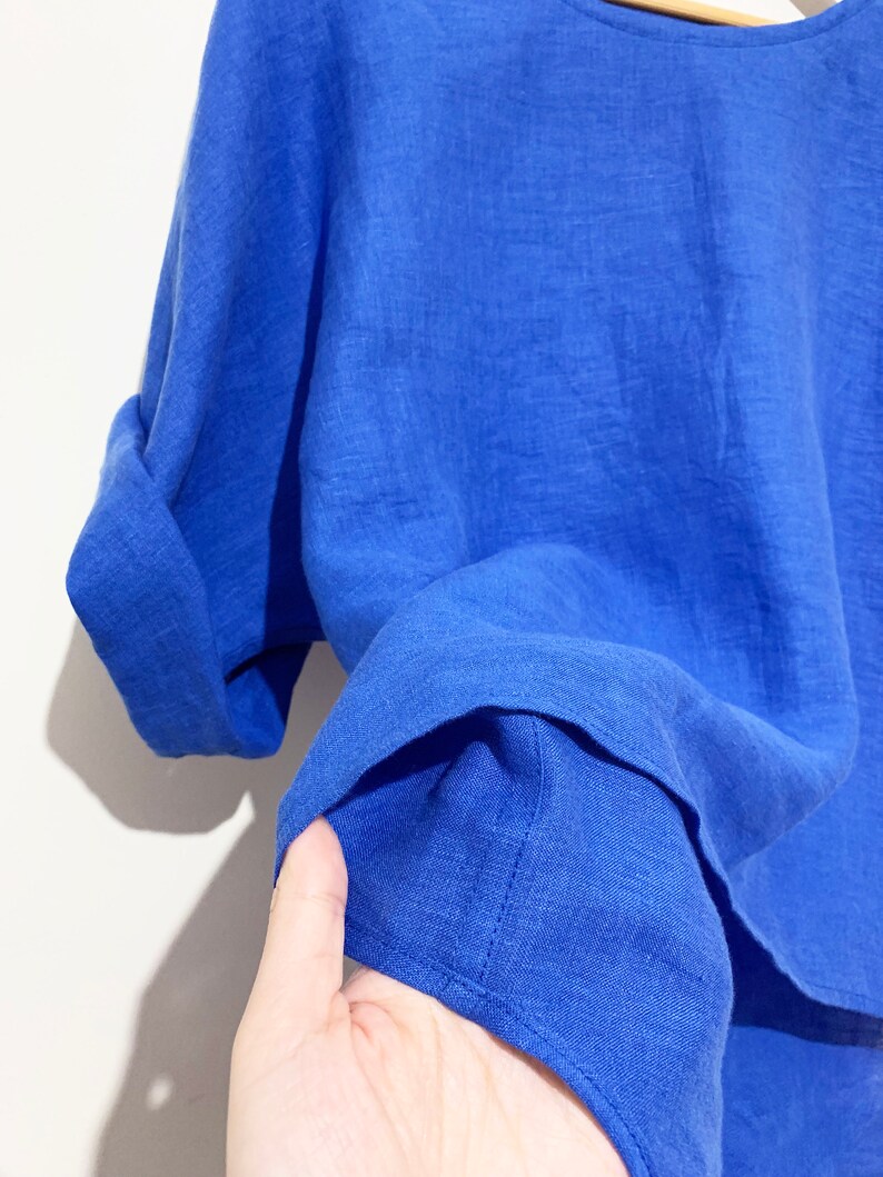 May include: A close-up of a vibrant blue linen top. The short-sleeved shirt has rolled-up cuffs and a round neckline. The fabric appears lightweight and slightly textured, suggesting breathability. The bright color makes it a stylish garment.