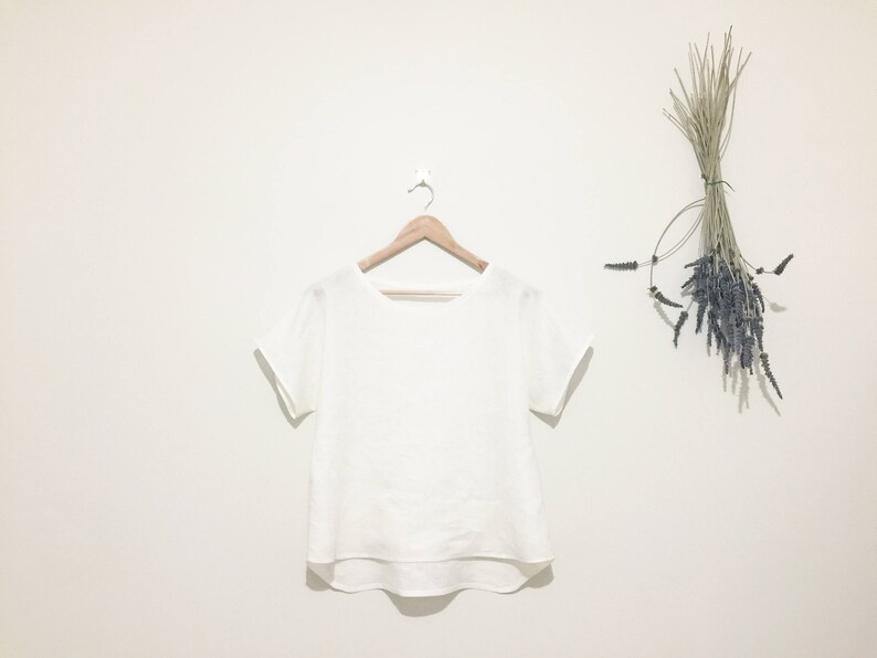 May include: A white, short-sleeved top hangs on a wooden hanger against a white wall. The top has a rounded neckline and a slightly curved hem. Dried lavender and grasses are tied together and hang on the wall to the right.