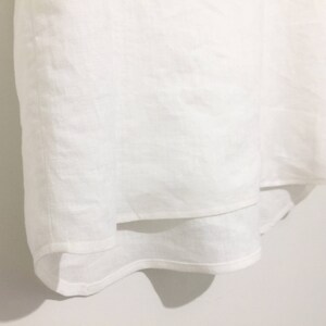 May include: A white linen garment with a layered design. The fabric appears to be lightweight and slightly textured. The garment is hanging against a white wall, with shadows adding depth.