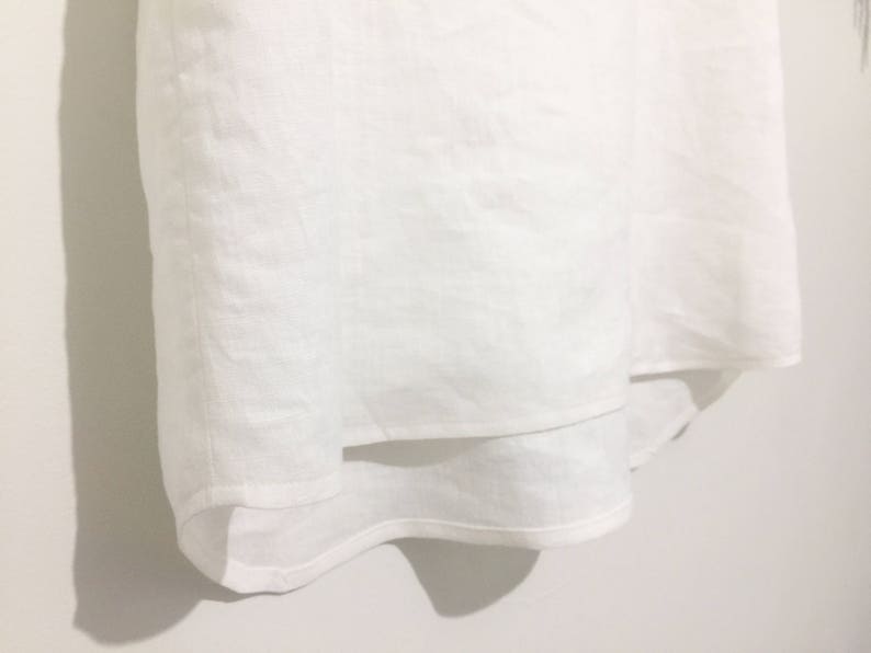 May include: A white linen garment with a layered design. The fabric appears to be lightweight and slightly textured. The garment is hanging against a white wall, with shadows adding depth.