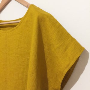 May include: Mustard yellow linen top with short sleeves, hanging on a wooden hanger. The top has a round neckline and a loose, flowing fit. The fabric appears to be lightweight and breathable, perfect for warm weather.