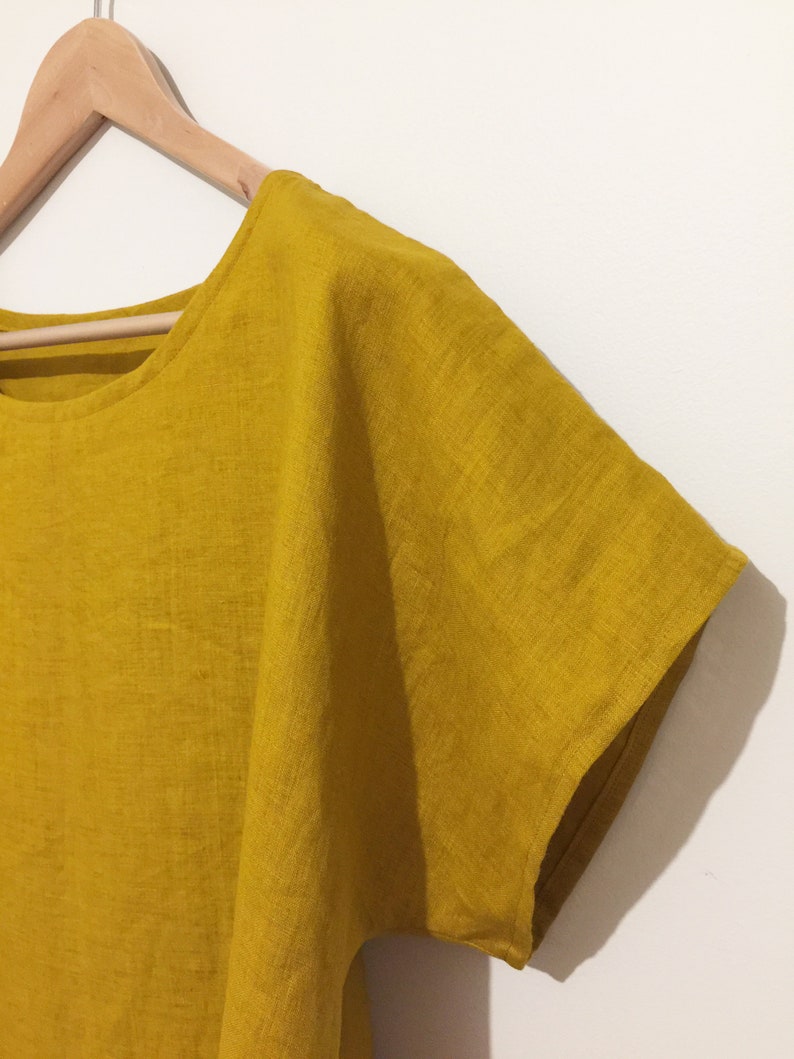 May include: Mustard yellow linen top with short sleeves, hanging on a wooden hanger. The top has a round neckline and a loose, flowing fit. The fabric appears to be lightweight and breathable, perfect for warm weather.