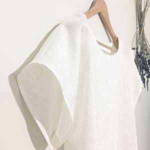 May include: A white linen top with short, wide sleeves hangs on a wooden hanger. The top has a simple, round neckline and a loose, flowing fit. The fabric appears to be lightweight and breathable. A bundle of dried flowers is visible in the background.