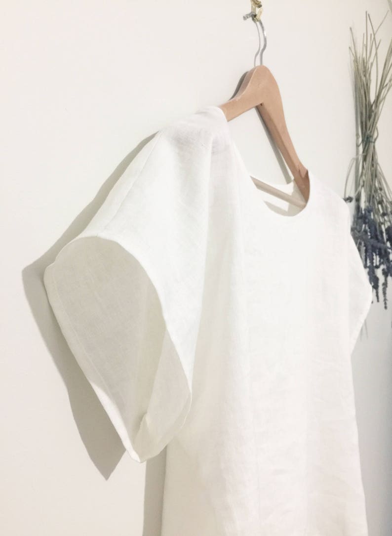 May include: A white linen top with short, wide sleeves hangs on a wooden hanger. The top has a simple, round neckline and a loose, flowing fit. The fabric appears to be lightweight and breathable. A bundle of dried flowers is visible in the background.