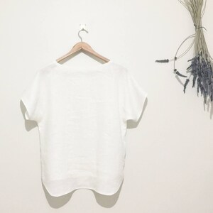 May include: A white linen top with short sleeves hangs on a wooden hanger against a white wall. A bundle of dried lavender is suspended on the right side. The top has a simple, relaxed design.