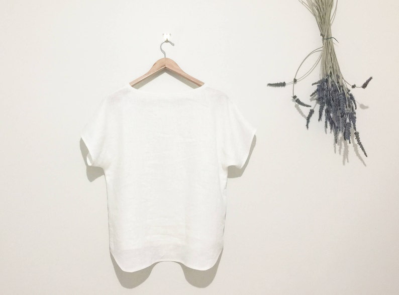 May include: A white linen top with short sleeves hangs on a wooden hanger against a white wall. A bundle of dried lavender is suspended on the right side. The top has a simple, relaxed design.