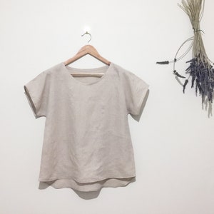 May include: A beige linen top with short sleeves hangs on a wooden hanger. The top has a rounded neckline and a slightly curved hem. A bundle of dried lavender and grasses hangs to the right.
