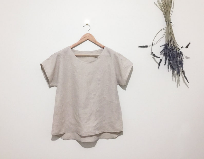 May include: A beige linen top with short sleeves hangs on a wooden hanger. The top has a rounded neckline and a slightly curved hem. A bundle of dried lavender and grasses hangs to the right.