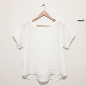 May include: A white, short-sleeved top with a rounded neckline hangs from a wooden hanger. The shirt has a loose fit and a slightly curved hemline. The background is a plain white wall, with a sprig of dried flowers to the right.