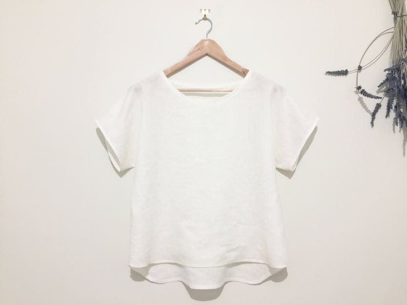 May include: A white, short-sleeved top with a rounded neckline hangs from a wooden hanger. The shirt has a loose fit and a slightly curved hemline. The background is a plain white wall, with a sprig of dried flowers to the right.