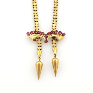 May include: A gold-toned necklace with a symmetrical design. The necklace features a series of small, round links and is adorned with red gemstones. The design includes two pendant-like elements with cone-shaped drops.