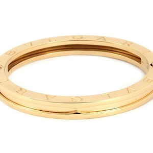 May include: A gold-colored bangle bracelet with the word "BVLGARI" engraved around the outer surface. The bracelet has a hinged design and a smooth, polished finish. The bracelet is approximately 2.5 inches in diameter.