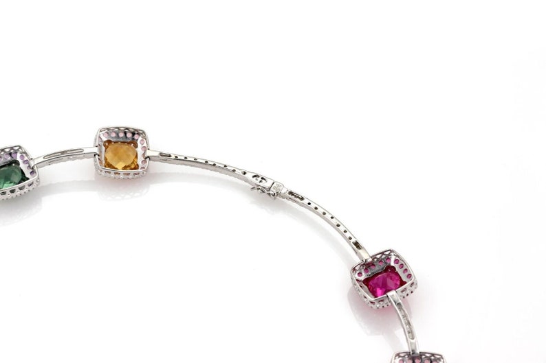 May include: A silver bracelet with square-cut gemstones in shades of yellow, pink, and green. The gemstones are set in a silver frame with small, clear stones around the edges. The bracelet is on a white background.