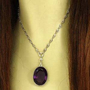 May include: A silver necklace with an oval, deep purple gemstone pendant. The chain has a delicate design, and the pendant is set in a silver frame. The necklace is displayed on a mannequin.
