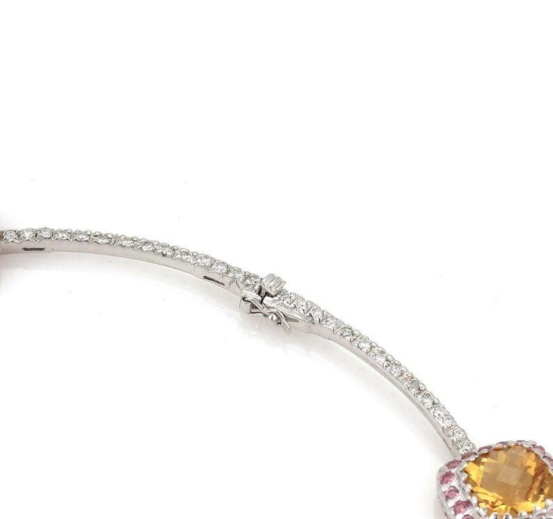 May include: A silver necklace with a clasp, featuring a large, faceted, yellow gemstone surrounded by small pink gemstones. The necklace is set with numerous small, clear gemstones.