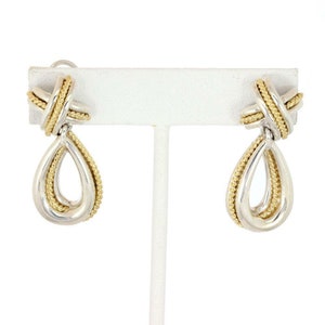May include: A pair of silver and gold-tone drop earrings. The earrings feature a teardrop shape with a gold-tone rope design around the edges and a gold-tone knot at the top. The earrings are displayed on a white stand.