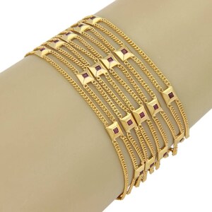 May include: Gold-toned bracelet featuring multiple strands of delicate chain. Each strand is connected by square-shaped links, each set with a small, square, red gemstone. The bracelet is shown on a wrist.