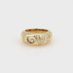 May include: A gold ring with the word "LOVE" spelled out in capital letters. The letters are encrusted with small, clear gemstones. A heart-shaped cutout with a single gemstone is visible within the "O".