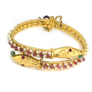 May include: A gold snake bangle bracelet with pink and white beads, green and red gemstones, and a hinged clasp.