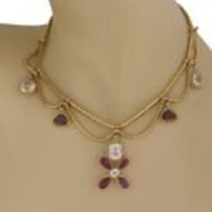 May include: A gold-toned necklace with a delicate design. The necklace features a series of small, clear and purple gemstones. The central pendant is a flower shape with a clear center stone and purple petals. The necklace is displayed on a mannequin.