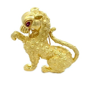 May include: A gold-colored brooch in the shape of a stylized lion. The lion has a textured, bubbled surface and a red gemstone eye. The brooch is posed in a dynamic stance, with one paw raised and its mouth open, revealing teeth.