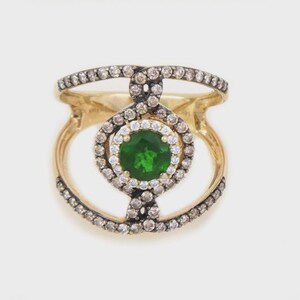 May include: A gold ring featuring a central round green gemstone surrounded by a halo of small white stones. The band is split into two open sections, both encrusted with numerous small brown stones that extend to the central setting.