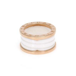 May include: A gold-tone ring with a white ceramic band. The ring features a circular design with the brand name engraved around the top edge. The white band is composed of several horizontal sections.