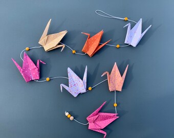 Może przedstawiać: Girlanda z kolorowych papierowych żurawi origami nawleczonych na żółte koraliki. Żurawie są w odcieniach różowego, pomarańczowego i fioletowego.