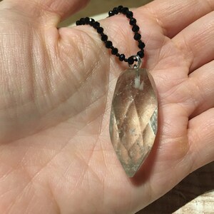 May include: A clear, faceted crystal pendant, teardrop-shaped, suspended from a black beaded necklace. The crystal has a light brown hue and is attached by a silver-coloured bail. The necklace is held in a hand.