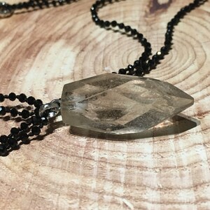 May include: A clear, faceted crystal pendant suspended from a black beaded chain. The crystal has a geometric shape and a translucent appearance. The necklace is presented against a wooden backdrop, offering a natural contrast.